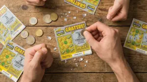 Mani che grattano un biglietto gratta e vinci su un tavolo di legno, con monete e frammenti di patina argentata