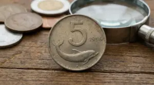 Moneta italiana da 5 lire con delfino e data 1956 su tavolo di legno, con lente d’ingrandimento sullo sfondo
