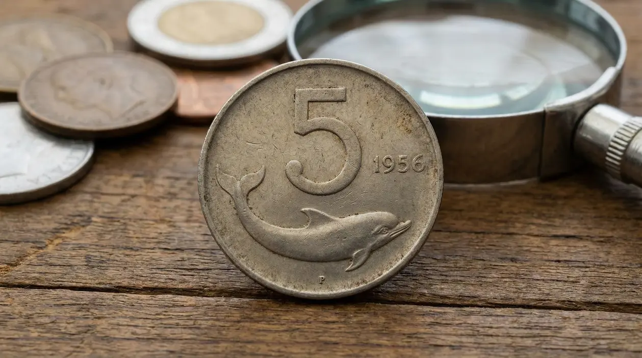 Moneta italiana da 5 lire con delfino e data 1956 su tavolo di legno, con lente d’ingrandimento sullo sfondo