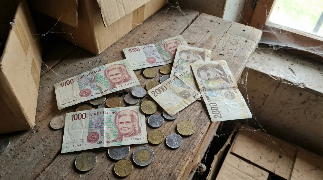 Vecchie banconote e monete in lire italiane su un tavolo polveroso in soffitta, accanto a scatole di cartone